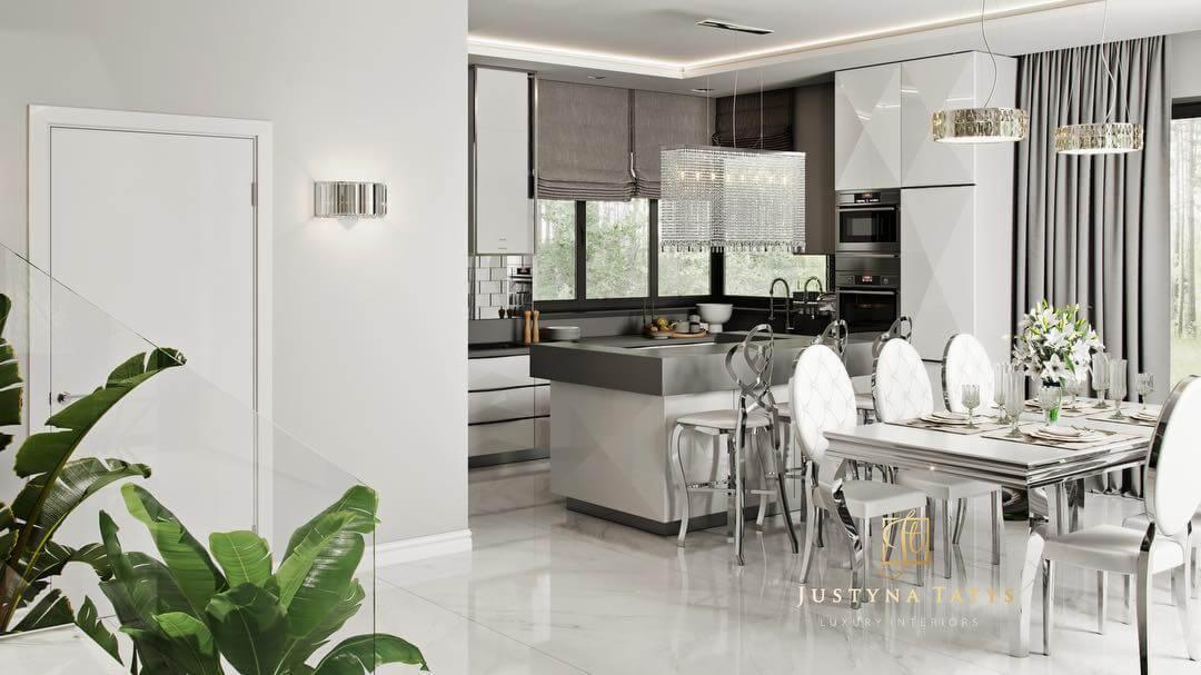 Nowoczesna, luksusowa kuchnia z wyspą i jadalnią, utrzymana w bieli i szarościach, z kryształowymi żyrandolami i rośliną doniczkową w rogu, widoczna ściana z oświetleniem.