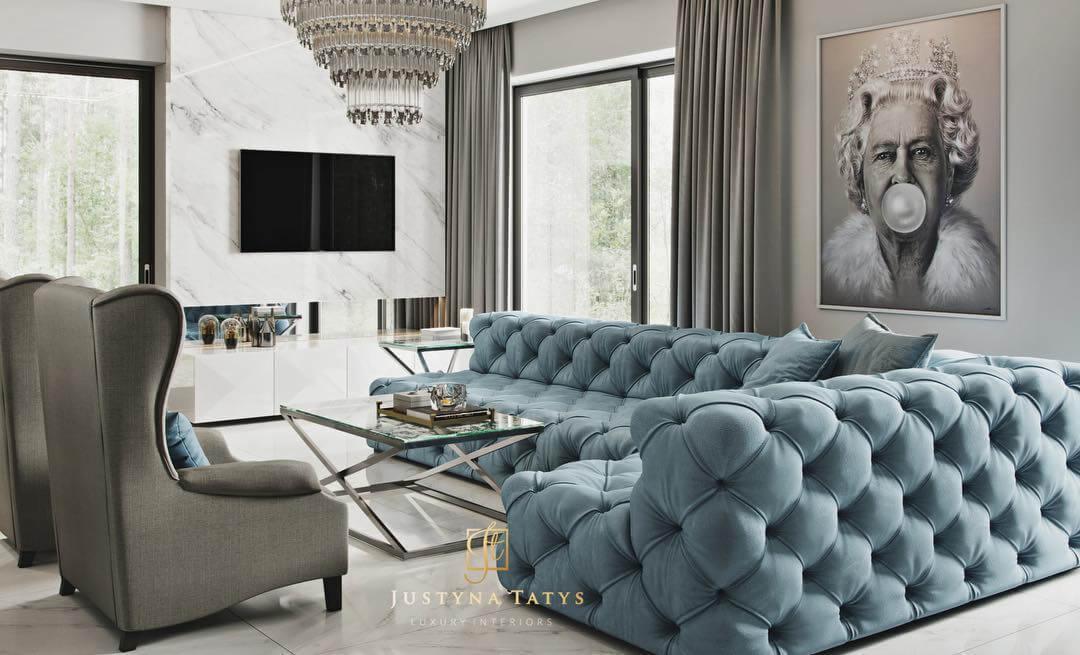 Luksusowy salon z pikowaną sofą w odcieniach błękitu, szarym fotelem typu uszak, marmurową ścianą z telewizorem, kryształowym żyrandolem i portretem królowej z gumą balonową.