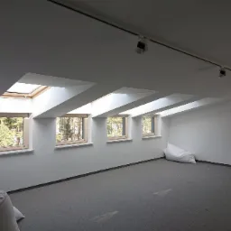 Poddasze z trzema oknami dachowymi w drewnianych ramach, szarym dywanem i workami z materiałem izolacyjnym w rogu, oświetlenie szynowe na suficie.