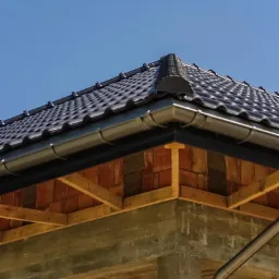 Detale wykończenia dachu z ciemnej dachówki ceramicznej i szarych rynien, widoczne elementy konstrukcyjne i mur, na tle błękitnego nieba. Kąt ujęcia od dołu.