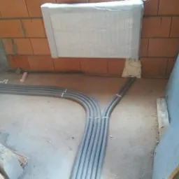 Szare rury instalacyjne prowadzone po podłodze w surowym stanie, widoczne cegły i ocieplenie, Wschowa.