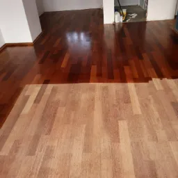 Fragment podłogi w trakcie renowacji: dolna część z surowego drewna, górna część po nałożeniu lakieru w odcieniu ciemnego brązu.