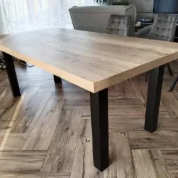 Prostokątny blat stołu z jasnego drewna, wsparty na czterech czarnych, kwadratowych nogach, na tle parkietu ułożonego w jodełkę.