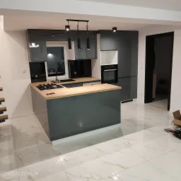 Nowoczesna kuchnia z szarymi, lakierowanymi frontami, drewnianym blatem, wyspą kuchenną i czarnymi lampami wiszącymi, widok na fragment schodów i rozpakowane narzędzia na podłodze.