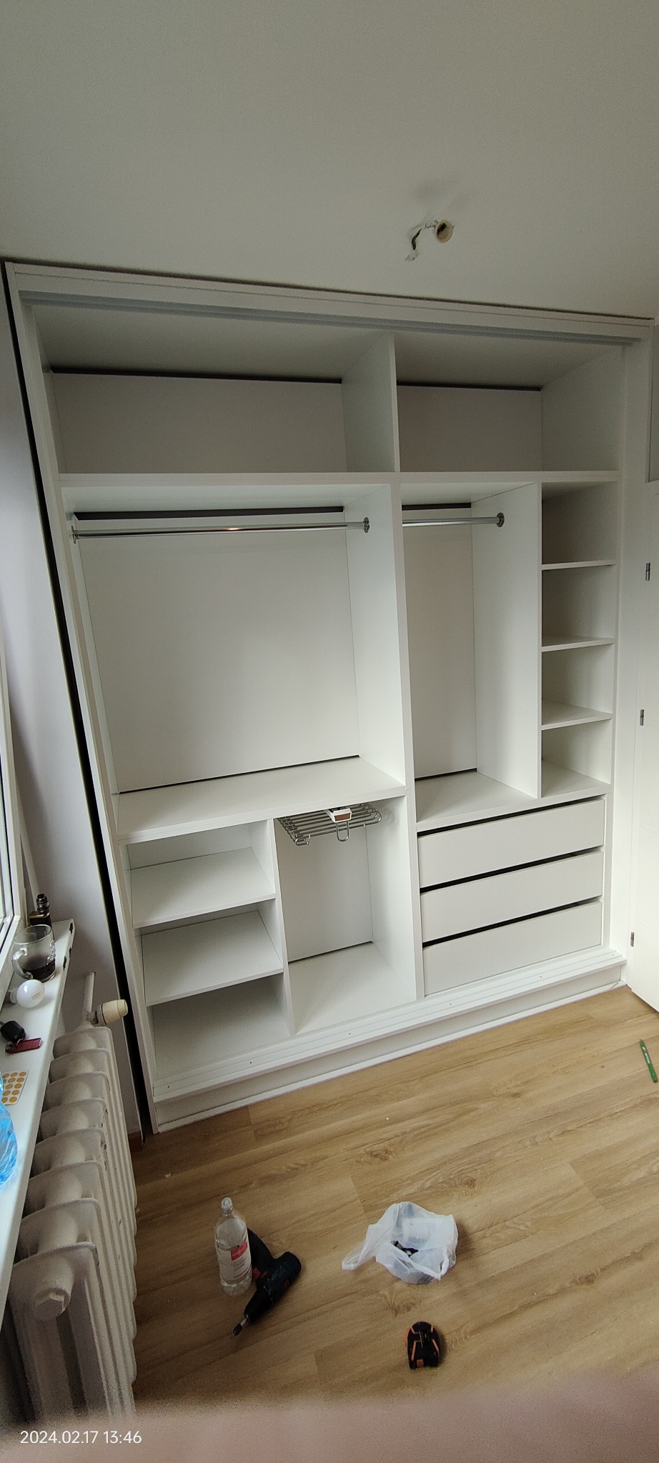 Biała szafa wnękowa z przesuwnymi drzwiami, widoczne półki, szuflady i drążki na ubrania. Na podłodze narzędzia i butelka. Minimalistyczny design.