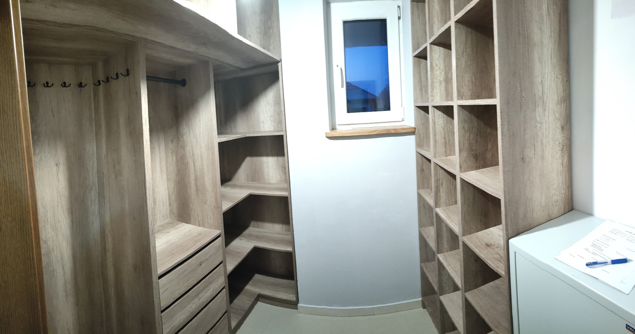 Jasna, drewnopodobna szafa wnękowa z półkami i szufladami, wkomponowana we wnękę z oknem. Widoczny fragment sejfu z dokumentami i długopisem.