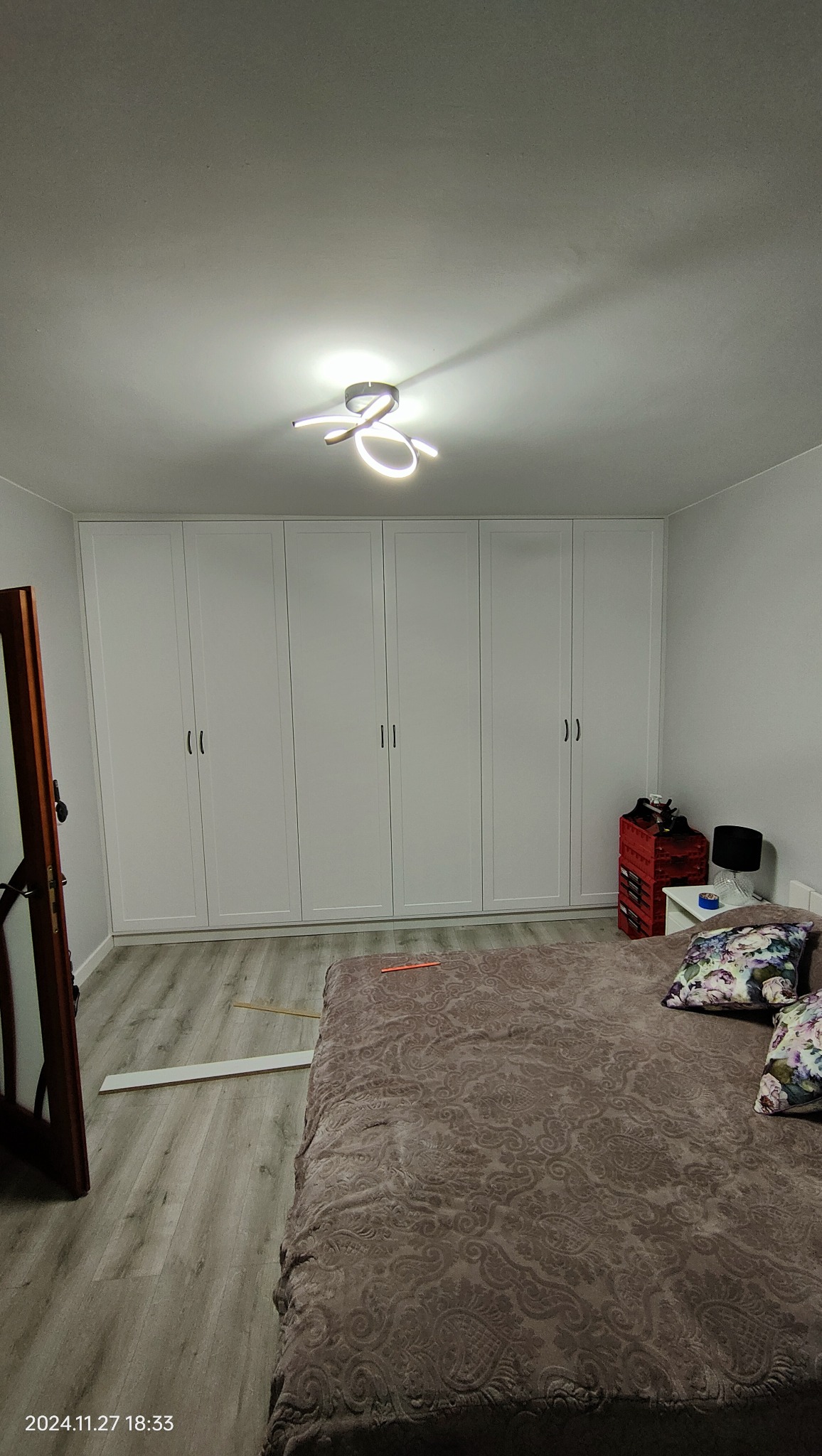 Biała szafa wnękowa z sześcioma drzwiami w sypialni, widok z perspektywy łóżka. Na podłodze panele, na suficie nowoczesna lampa.