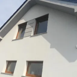 Fragment białej elewacji domu dwukondygnacyjnego z drewnianymi oknami i szarym dachem, widok z dołu na ścianę szczytową z fragmentem dekoracyjnego panelu elewacyjnego między oknami na poddaszu.