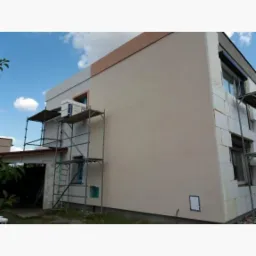 Dwupiętrowy dom w trakcie ocieplania, z widocznym rusztowaniem, częściowo otynkowaną elewacją w beżowym kolorze oraz fragmentami białego styropianu na ścianach.