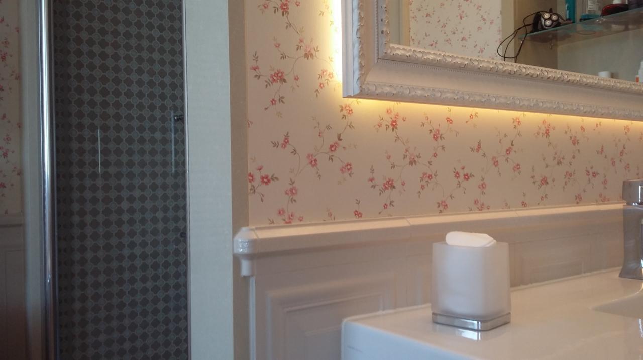 Umywalka z białym blatem, bateria i pojemnik na chusteczki, w tle kabina prysznicowa z wzorzystą szybą i ściana w kwiatowy wzór z podświetlanym lustrem.