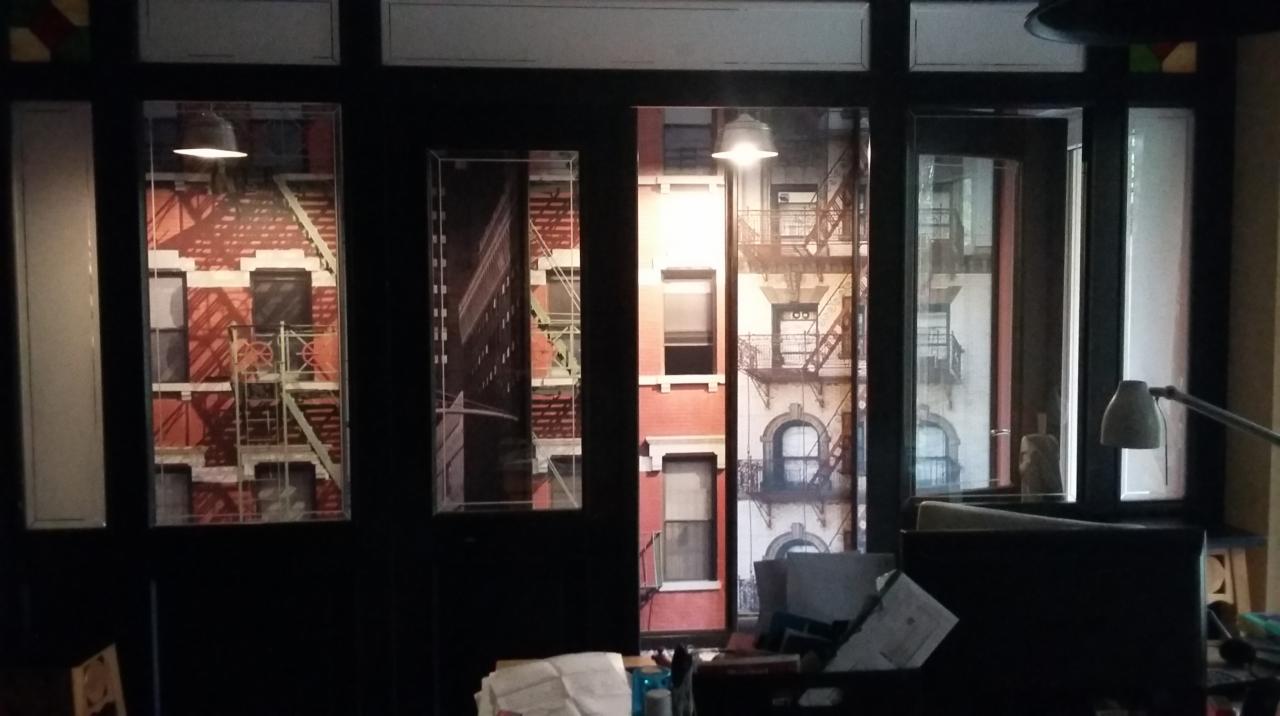 Biuro z widokiem na fototapetę przedstawiającą nowojorskie kamienice z pożarowymi schodami, oświetlone lampą uliczną, widoczne biurko z lampką.