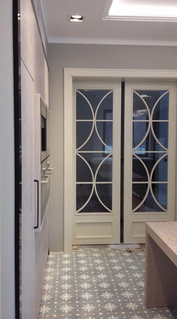 Jasna kuchnia z zabudową meblową, wbudowanym piekarnikiem i dekoracyjnymi przeszklonymi drzwiami przesuwnymi z motywem geometrycznym, oraz wzorzystą podłogą w odcieniach szarości i bieli.