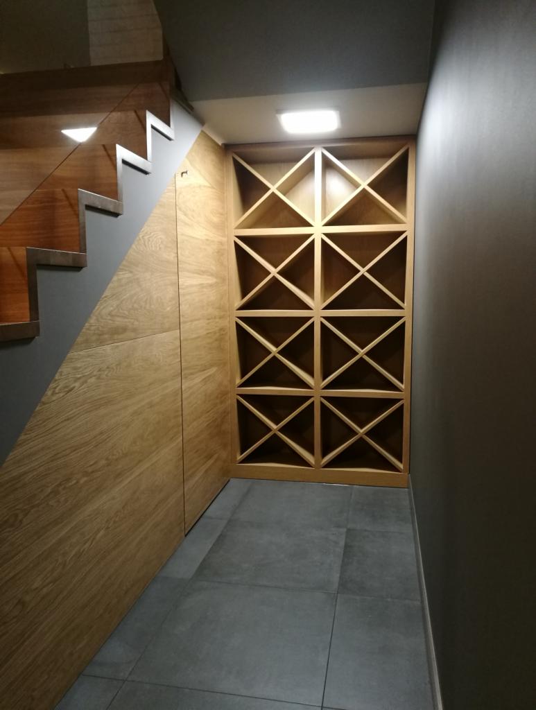 Drewniana zabudowa pod schodami z regałem na wino o geometrycznym wzorze, szare płytki podłogowe, minimalistyczne oświetlenie sufitowe.