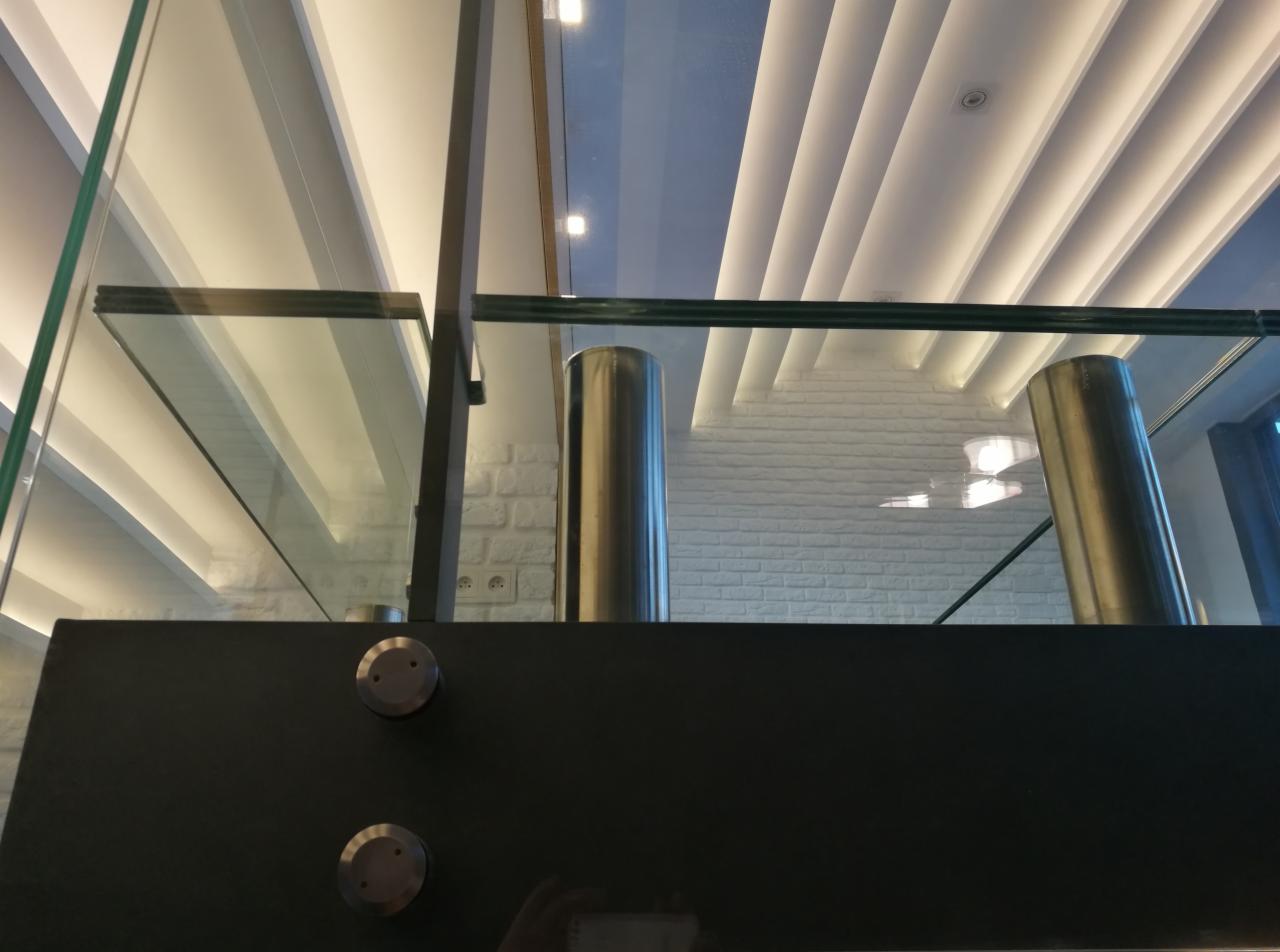 Szklana balustrada z metalowymi słupkami i czarnym mocowaniem, widok z dołu na sufit z podłużnym oświetleniem LED i białą ceglaną ścianę w tle.