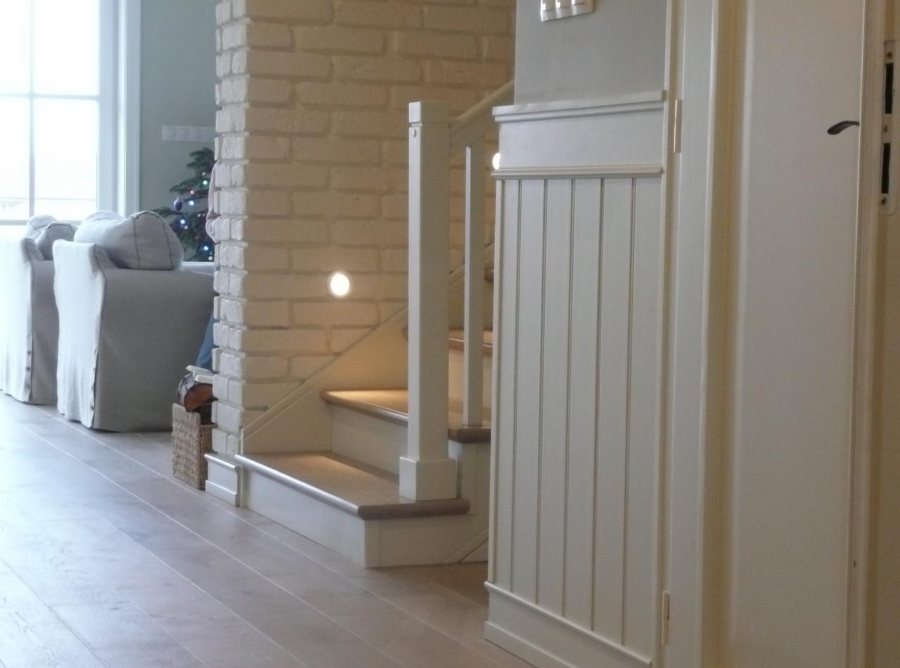 Jasne drewniane schody z podświetleniem w ścianie z cegły, obok biała szafa w stylu klasycznym i widoczne sofy w tle.