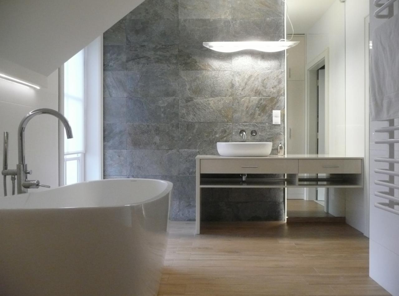 Nowoczesna łazienka z wolnostojącą wanną, umywalką nablatową na szafce z szufladami oraz ścianą wyłożoną szarym kamieniem naturalnym pod skośnym sufitem.