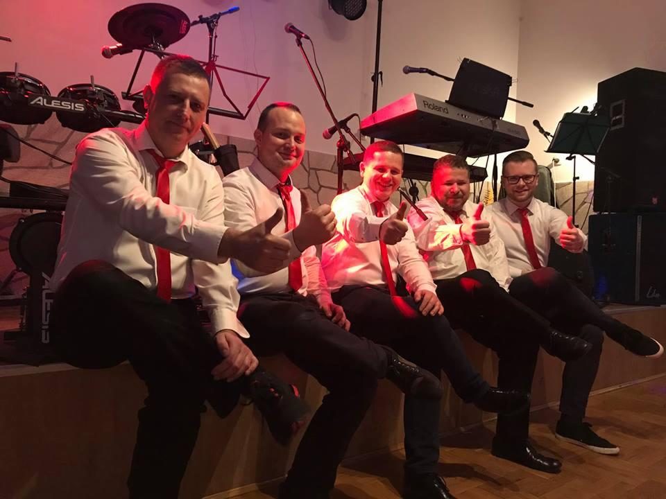 Pięciu mężczyzn w białych koszulach i czerwonych krawatach, siedzących na scenie z instrumentami muzycznymi w tle, kciuki w górę, Wieliczka.