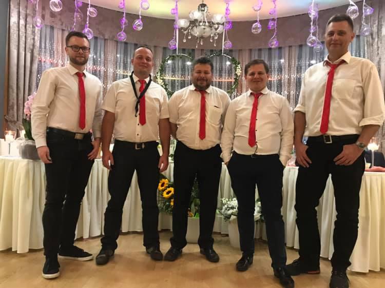 Pięcioosobowy zespół muzyczny w białych koszulach i czerwonych krawatach pozuje na tle dekoracji sali weselnej z kwiatami i lampionami.