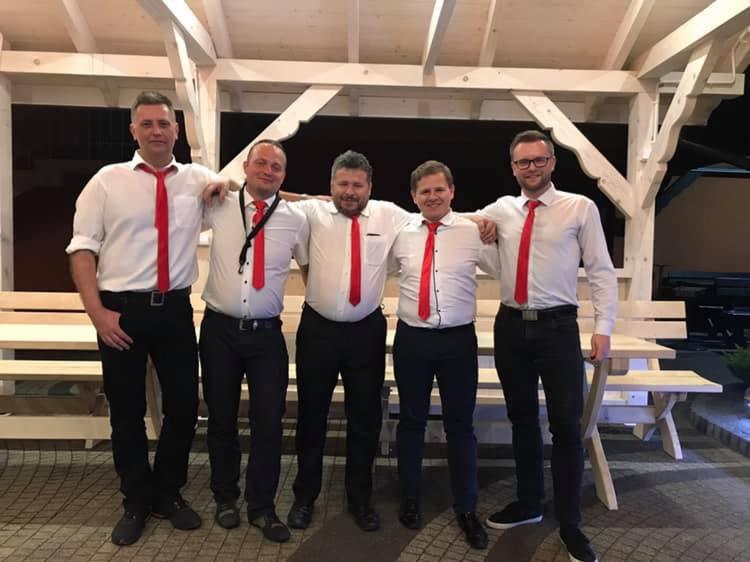 Pięcioosobowy zespół w białych koszulach i czerwonych krawatach, pozujący pod drewnianą altaną z ławkami, jeden z muzyków ma czarną smycz na szyi.