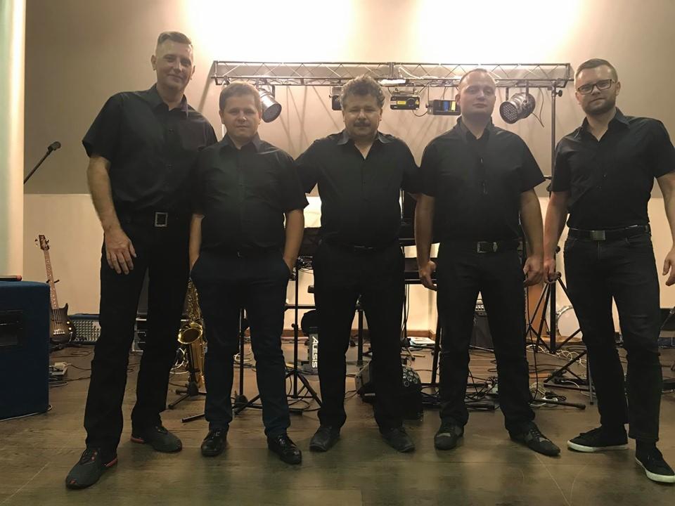 Pięcioosobowy zespół muzyczny w czarnych koszulach pozuje na scenie przed sprzętem nagłośnieniowym, w tle widoczny saksofon i gitara basowa.