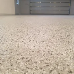 Jasna, błyszcząca posadzka z żywicy z widocznymi drobnymi, nieregularnymi płatkami w odcieniach bieli i szarości, na tle szarej ściany i zamkniętej bramy garażowej.