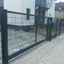 Nowoczesne, przesuwne, grafitowe ogrodzenie panelowe z widoczną furtką, zamontowane na posesji z kostki brukowej, na tle budynku mieszkalnego.