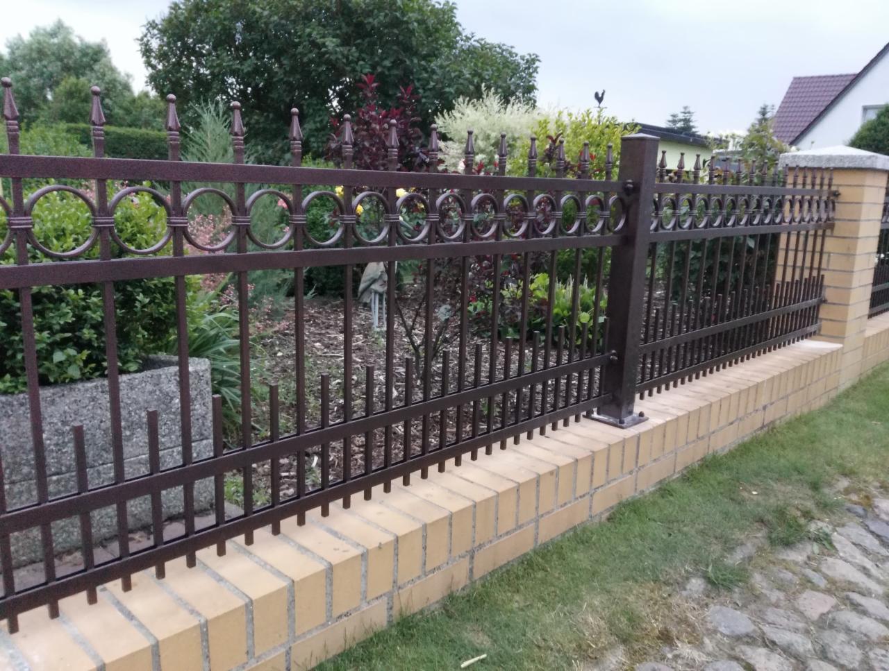 Ciemnobrązowe ogrodzenie z metalowymi prętami i okręgami na ceglanym murku, widok na zadbany ogród z różnorodną roślinnością.