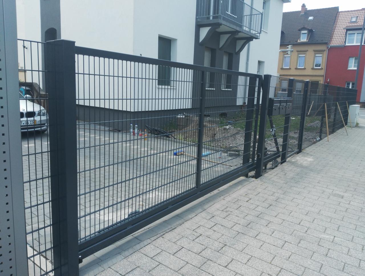 Nowoczesne, przesuwne, grafitowe ogrodzenie panelowe z widoczną furtką, zamontowane na posesji z kostki brukowej, na tle budynku mieszkalnego.