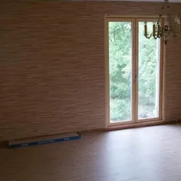 Pokój z panelami na podłodze i ścianach, okno z widokiem na zieleń, wiszący żyrandol i paczka paneli przy ścianie.