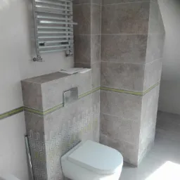 Szara łazienka z podwieszaną toaletą, grzejnikiem drabinkowym i geometrycznym wzorem na zabudowie stelaża, wykończona płytkami imitującymi kamień z zielonym akcentem.