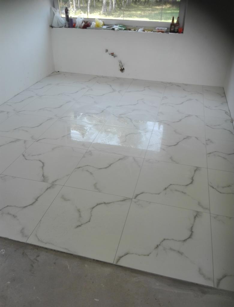 Wyłożona białymi płytkami podłoga z szarym wzorem marmuru w pomieszczeniu z oknem, przez które widać drzewa. Część podłogi jeszcze nie wykończona wylewką.