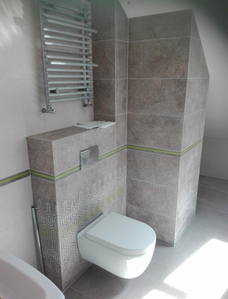 Szara łazienka z podwieszaną toaletą, grzejnikiem drabinkowym i geometrycznym wzorem na zabudowie stelaża, wykończona płytkami imitującymi kamień z zielonym akcentem.