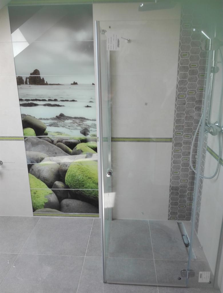 Nowoczesna kabina prysznicowa z szklanymi drzwiami, wyłożona płytkami z motywem kamieni pokrytych mchem i widokiem na morze w tle, uzupełniona mozaiką w kształcie plastra miodu.