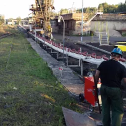 Spawacz w kasku przygotowuje się do spawania konstrukcji przenośnika taśmowego na terenie przemysłowym, widoczna spawarka na kółkach i blacha na ziemi.