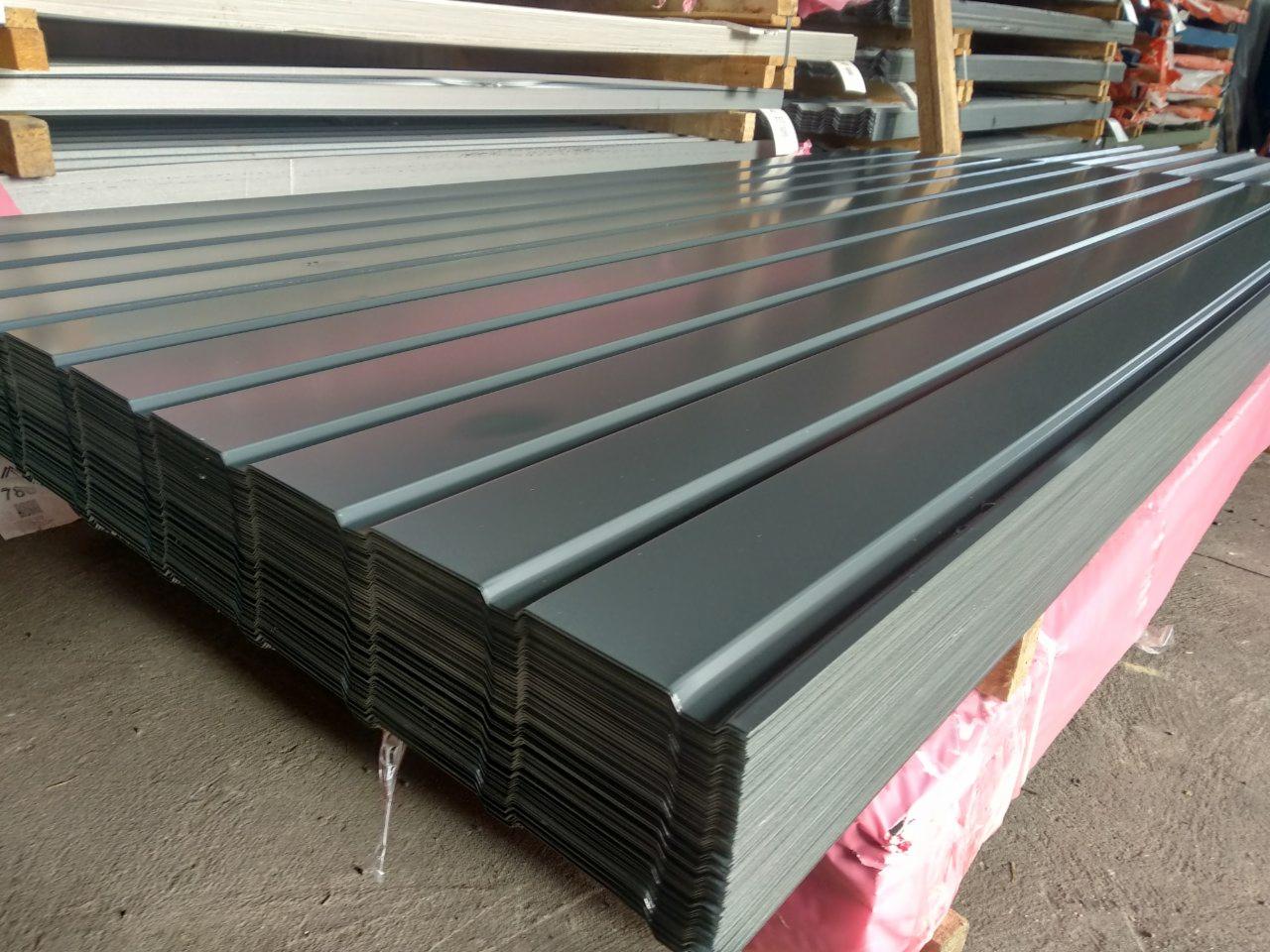 Stos arkuszy blachy trapezowej w kolorze grafitowym, ułożonych na palecie z różową folią ochronną, widoczne ślady transportu.