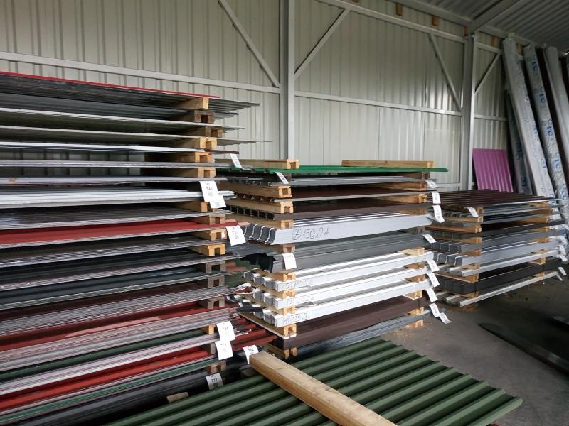 Magazyn z paletami różnokolorowej blachy trapezowej i profili metalowych pod zadaszeniem o konstrukcji stalowej.