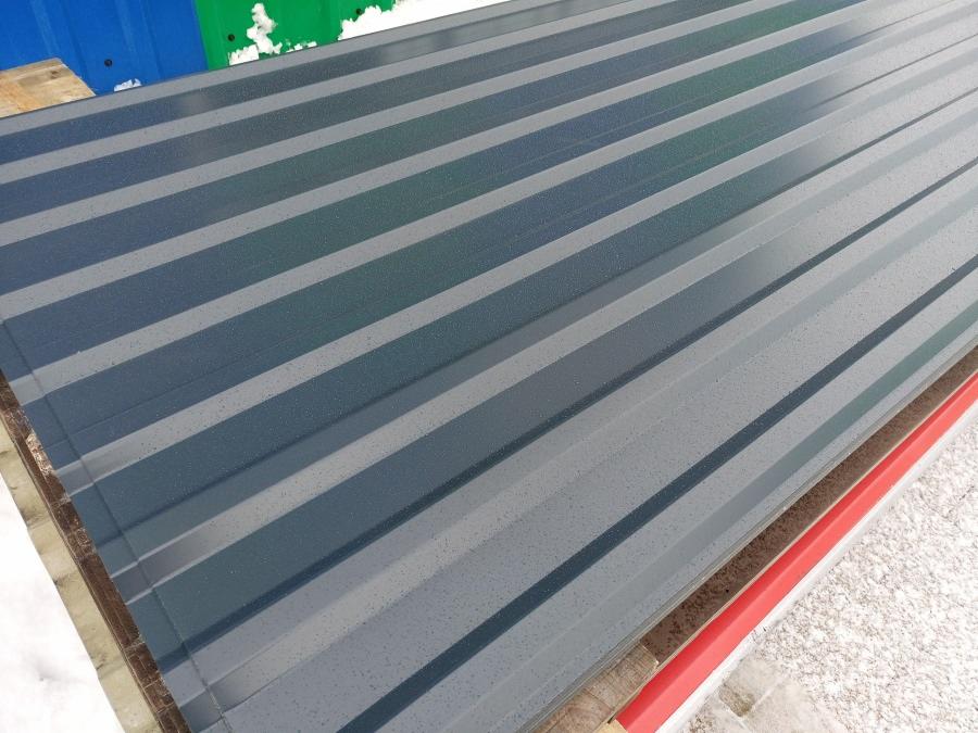 Stos arkuszy blachy trapezowej w kolorze grafitowym, ułożonych na drewnianych legarach z widocznymi kroplami wody na powierzchni.