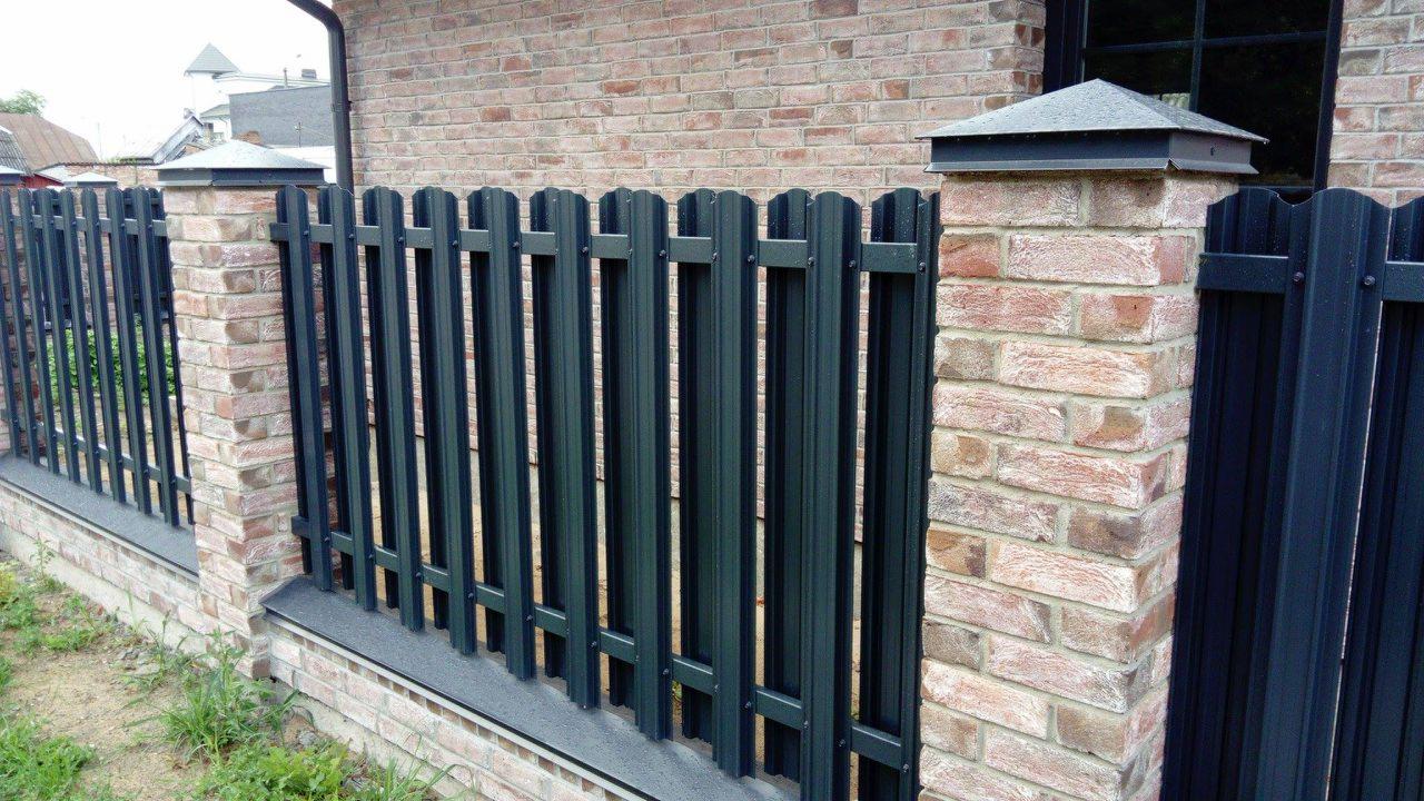 Nowoczesne, ciemnozielone ogrodzenie metalowe z pionowymi sztachetami, osadzone na niskim murku z cegły klinkierowej z szarą nakładką, widoczne z bliska na tle ceglanego domu.