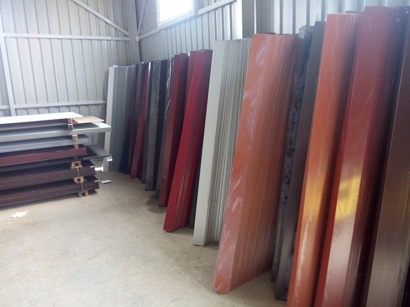 Magazyn blachy trapezowej w różnych kolorach, ułożonej pionowo, zabezpieczonej folią, widoczne arkusze szare i ułożone poziomo w lewym rogu.