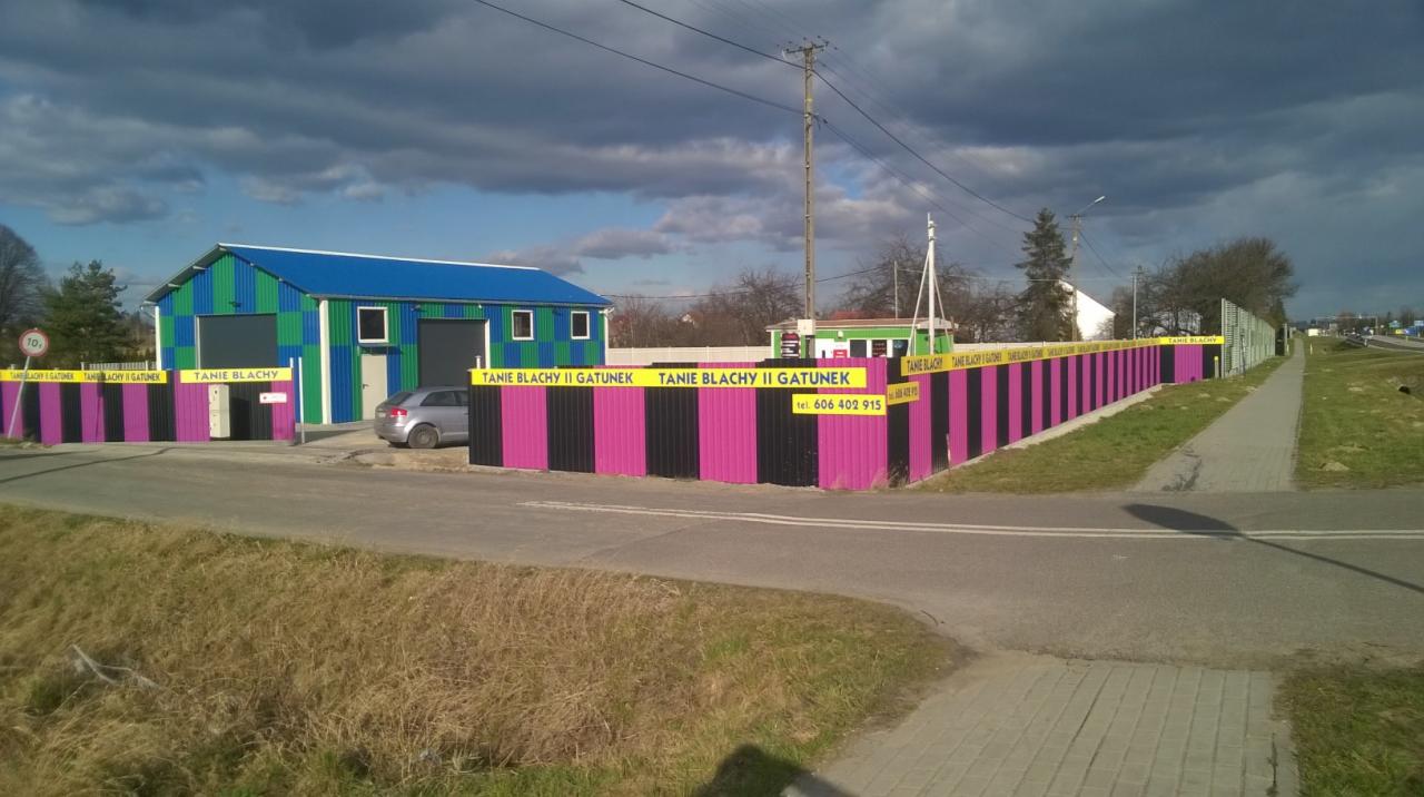 Magazyn z blachy trapezowej z niebiesko-zielonymi ścianami i ogrodzeniem w czarno-różowe pasy z reklamą 'Tanie Blachy II Gatunek' i numerem telefonu.