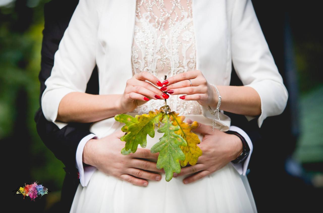 Panna młoda w białej sukni i bolerku trzyma gałązkę dębu z obrączkami, obejmowana przez pana młodego w czarnym garniturze. Czerwone paznokcie panny młodej i srebrna bransoletka dodają elegancji.