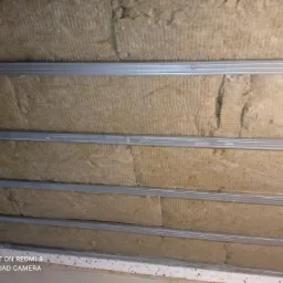 Wypełniona wełną mineralną konstrukcja sufitu podwieszanego, widoczne metalowe profile i fragment ściany poniżej.