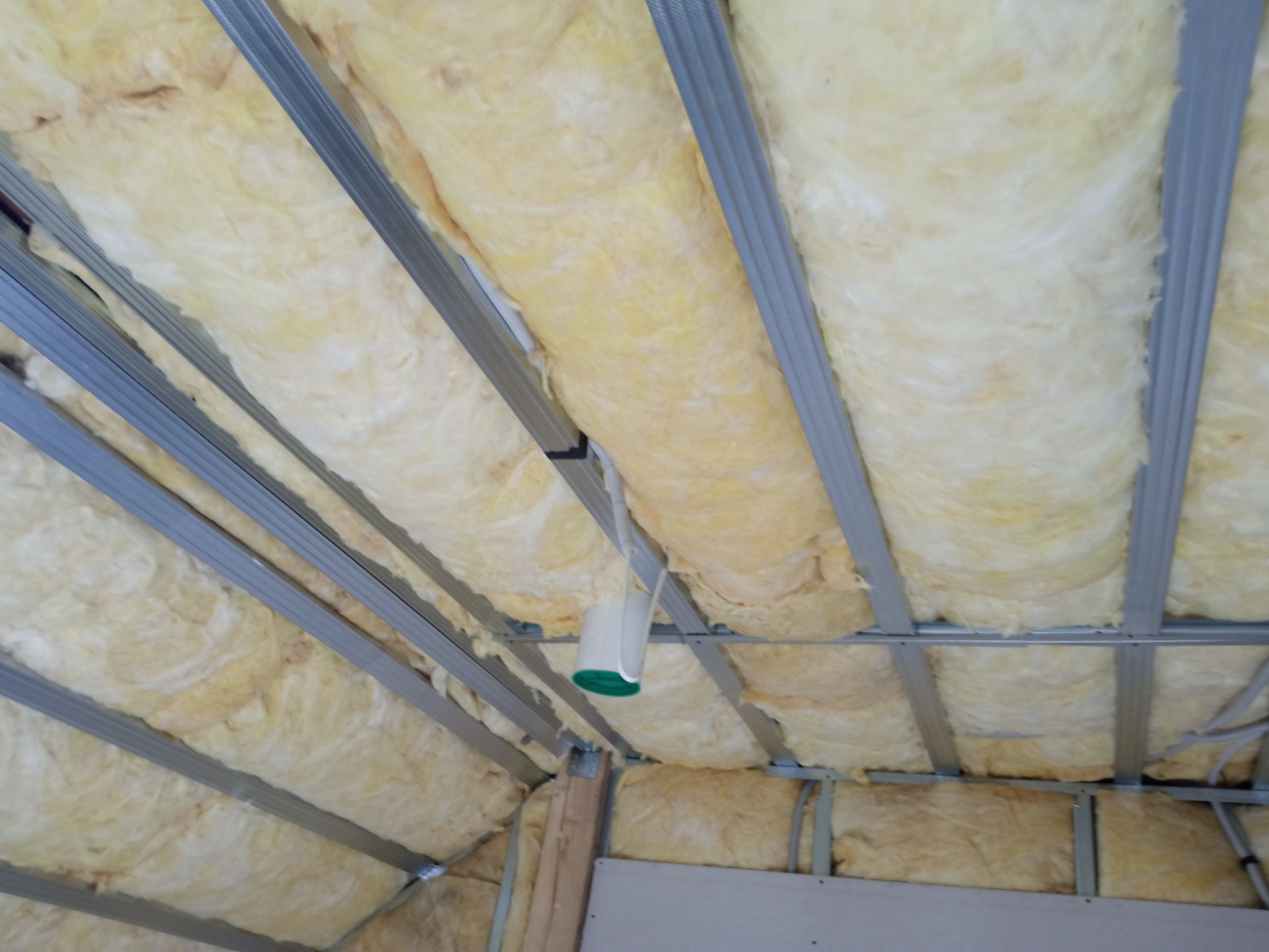 Wypełnienie wełną mineralną między metalowymi profilami konstrukcji poddasza, widoczne przewody elektryczne i fragment ściany z płyt gipsowo-kartonowych.