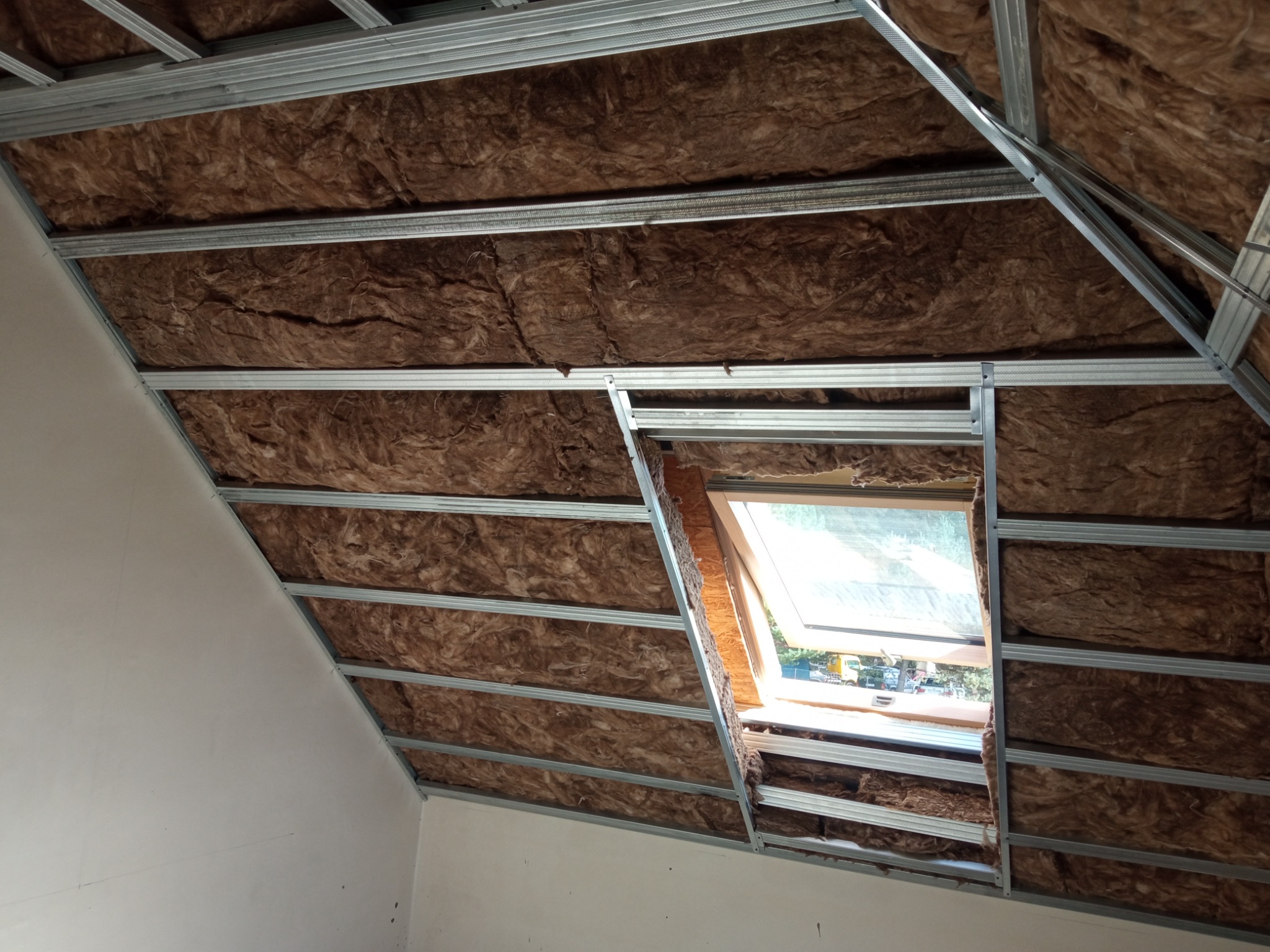 Skośne poddasze z oknem dachowym w trakcie izolacji brązową wełną mineralną, widoczna metalowa konstrukcja szkieletowa pod przyszłą zabudowę.