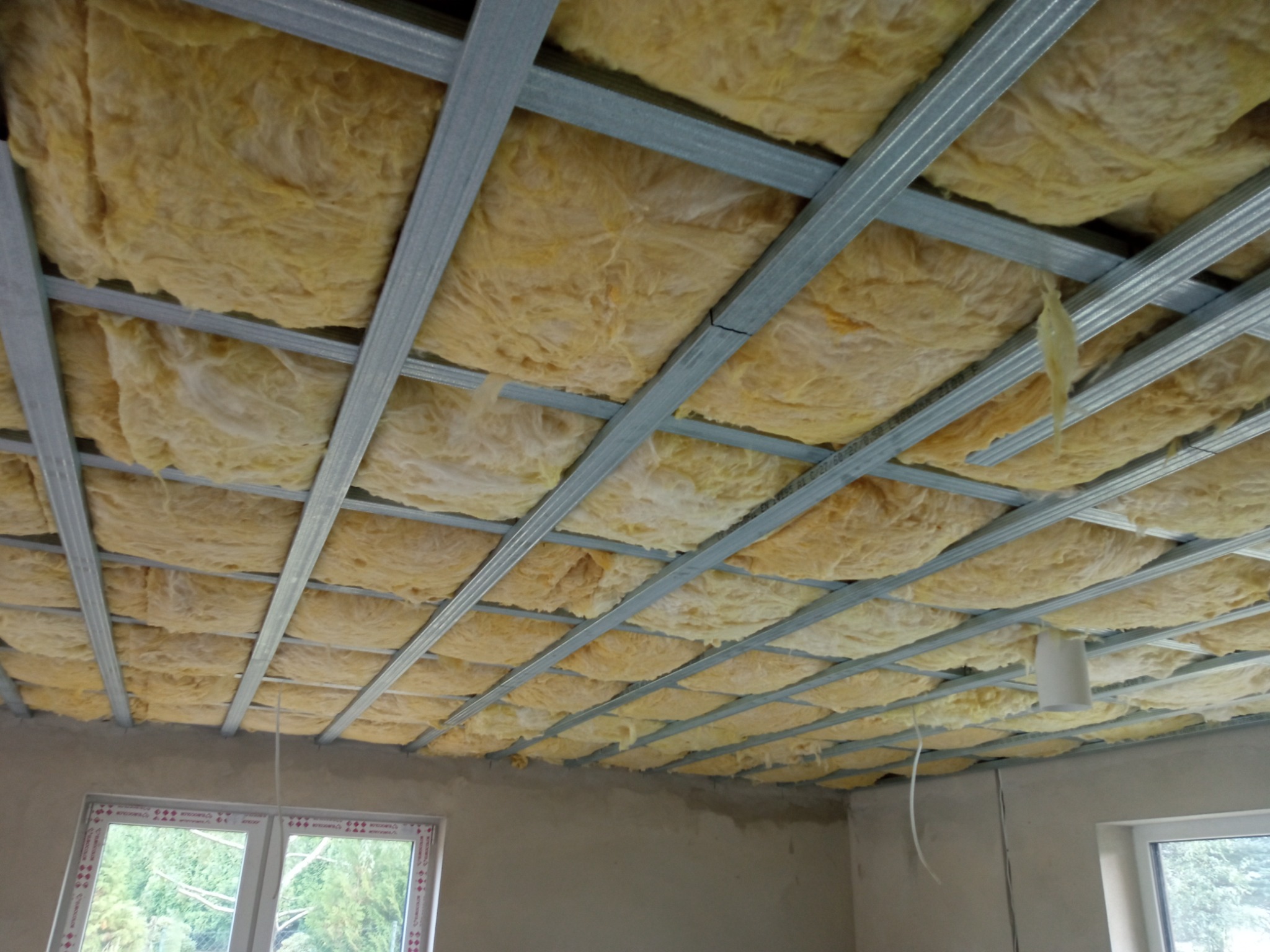 Montaż izolacji z wełny mineralnej w metalowym stelażu sufitowym, widok z dołu, pomieszczenie w stanie surowym, okna z widokiem na zieleń.