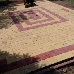 Układanie kostki brukowej w Rudzie Śląskiej z dekoracyjnym wzorem w kształcie kwadratu z czerwonej kostki na żółtym tle, widoczne narzędzia i fragmenty pracującej osoby.