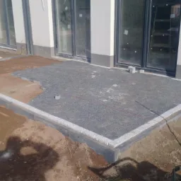 Budowa tarasu: częściowo ułożona nawierzchnia z drobnej, szarej kostki na siatce, obramowana jasnymi kostkami, widoczny przygotowany grunt i narzędzia, budynek z dużymi oknami w tle.