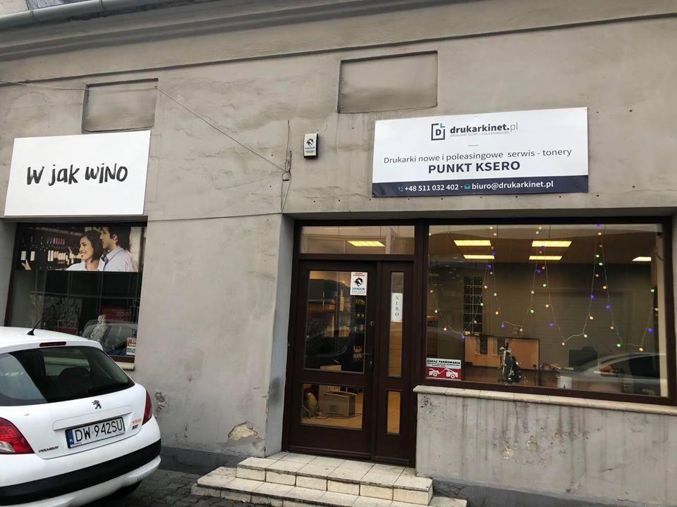 Fasada budynku z szyldem 'drukarkinet.pl' oferującego serwis drukarek, tonery i punkt ksero, obok wejścia do sklepu z winami 'W jak wino'.