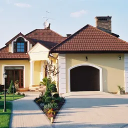 Żółty dom jednorodzinny z brązowym dachem, kolumnami przy wejściu, dwuskrzydłowymi drzwiami, garażem z bramą segmentową i zagospodarowanym ogrodem z chodnikiem z kostki.