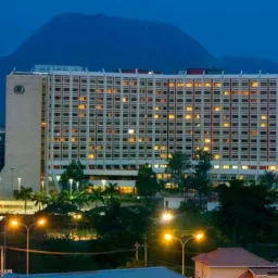 Wieczorny widok na wielopiętrowy budynek hotelowy z podświetlonym logo na dachu, na tle ciemnego nieba i zalesionych wzgórz.